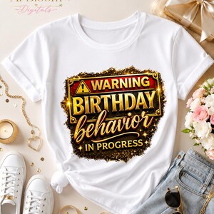 May include: A white short-sleeved t-shirt featuring a graphic design with a yellow warning triangle icon and the text "WARNING BIRTHDAY behavior IN PROGRESS" in bold gold and red lettering, set against a dark, textured, glittery background.