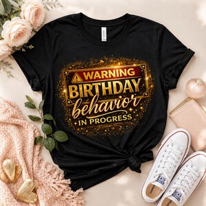 May include: A black short-sleeved t-shirt featuring a graphic print with a gold-bordered rectangular sign. The text reads WARNING BIRTHDAY behavior IN PROGRESS in gold and white lettering, surrounded by a sparkling gold glitter effect.