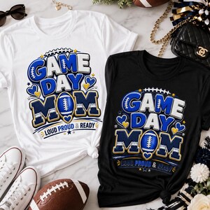 May include: A white t-shirt and a black t-shirt, each featuring a graphic design with the text "GAME DAY MOM" and "LOUD PROUD & READY" in blue, white, and gold, accented with football, heart, and star illustrations.