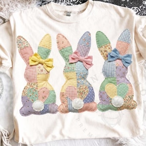 May include: A white t-shirt featuring three patchwork bunny rabbits. Each bunny has a different colored bow and is made of various patterned fabrics, including floral, polka dot, and gingham. The bunnies have white pom-pom tails.