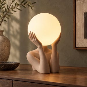 May include: A table lamp featuring a beige, textured sculpture of a torso and arms holding a large, spherical, glowing white glass shade. The light emits a warm, soft illumination from the center of the sphere, resting on a flat surface.
