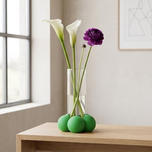 May include: A glass vase holding white calla lilies and a purple ranunculus. The vase has a green spherical base. The arrangement sits on a wooden table. The table is in front of a window.