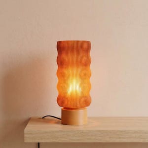 May include: A modern table lamp with a cylindrical, textured shade in shades of orange and brown. The lamp sits on a round, light-colored wooden base. The lamp is plugged into an electrical outlet.
