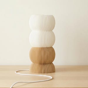 May include: A modern, stacked lamp with a unique design. The lamp features three sections: two white and one tan, all with vertical ribbed detailing. A white cord extends from the base. The lamp is on a wooden surface.