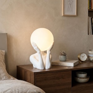 Può includere: Una lampada da tavolo scultorea bianca raffigurante una figura umana stilizzata con le mani sollevate a sostenere un grande paralume sferico in vetro illuminato. La lampada emette una luce calda e diffusa che esalta la finitura opaca liscia.