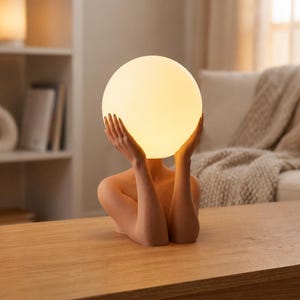 May include: A table lamp featuring a sculpted figure with arms raised to hold a glowing, spherical white glass shade. The base is a smooth, matte-finish human torso and head-less form in a neutral beige tone, supporting the illuminated orb.