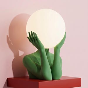 May include: A sculptural lamp featuring a green figure with raised arms holding a glowing white spherical light bulb. The figure is positioned on a flat red rectangular base. The text Serenity Sculptural Lamp in Green is printed below.