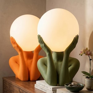 May include: Two table lamps featuring textured, matte-finish human-like busts in orange and olive green. Each bust has raised arms holding a large, spherical, glowing white glass shade. The lamps are positioned on top of stacked hardcover books.