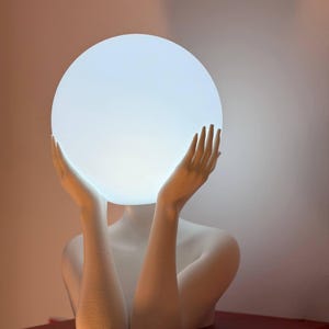 Puede incluir: Una l&aacute;mpara escult&oacute;rica blanca que presenta el busto de una figura con los brazos levantados. Las manos est&aacute;n colocadas a ambos lados de una gran bombilla esf&eacute;rica blanca y brillante que sirve como cabeza de la escultura.