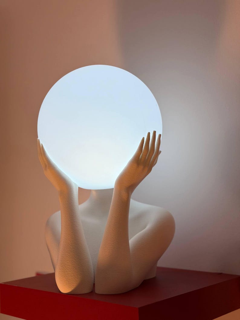 Puede incluir: Una l&aacute;mpara escult&oacute;rica blanca que presenta el busto de una figura con los brazos levantados. Las manos est&aacute;n colocadas a ambos lados de una gran bombilla esf&eacute;rica blanca y brillante que sirve como cabeza de la escultura.