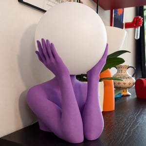Puede incluir: Una l&aacute;mpara de mesa con una escultura de color morado mate que representa un torso y brazos, con las manos ahuecadas alrededor de una gran pantalla esf&eacute;rica de cristal blanco. La escultura est&aacute; erguida, con las manos sosteniendo el globo.