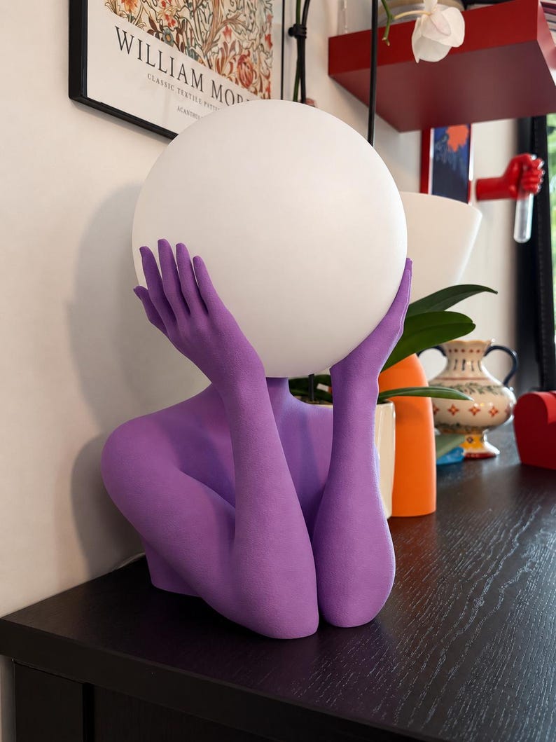 Puede incluir: Una l&aacute;mpara de mesa con una escultura de color morado mate que representa un torso y brazos, con las manos ahuecadas alrededor de una gran pantalla esf&eacute;rica de cristal blanco. La escultura est&aacute; erguida, con las manos sosteniendo el globo.