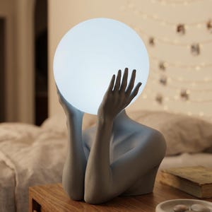 May include: A gray sculptural table lamp featuring a human torso and arms reaching upward to hold a large, glowing, spherical white light bulb. The lamp is crafted with a matte finish and depicts a figure with hands positioned around the orb.