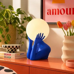 Puede incluir: Una l&aacute;mpara de mesa con una escultura de color azul brillante de un torso y manos que sostienen una gran pantalla esf&eacute;rica de cristal blanco brillante. La base de la l&aacute;mpara tiene forma de persona desde el pecho, con las manos levantadas.