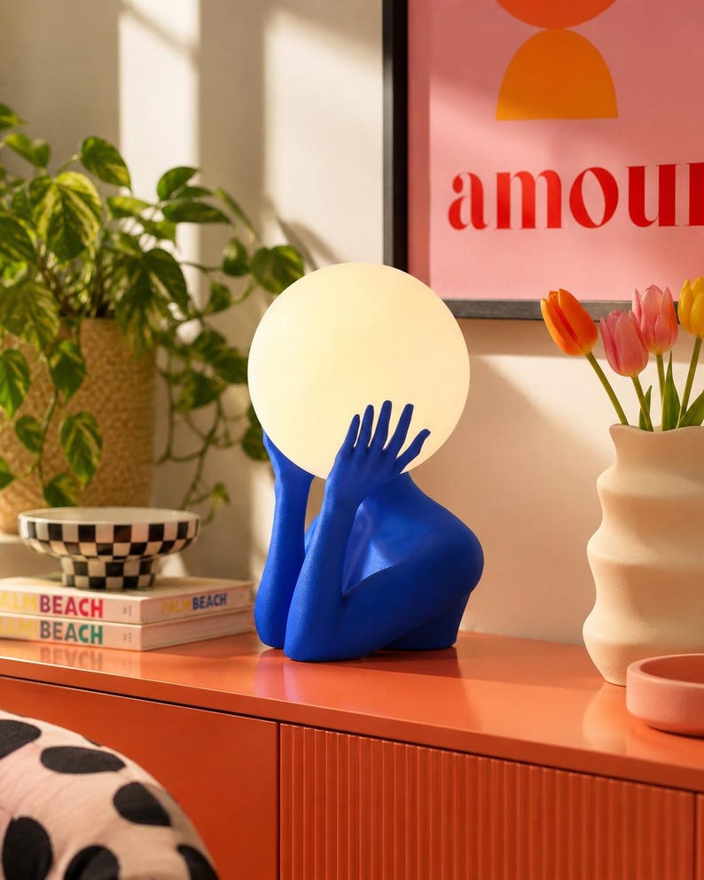 Puede incluir: Una l&aacute;mpara de mesa con una escultura de color azul brillante de un torso y manos que sostienen una gran pantalla esf&eacute;rica de cristal blanco brillante. La base de la l&aacute;mpara tiene forma de persona desde el pecho, con las manos levantadas.