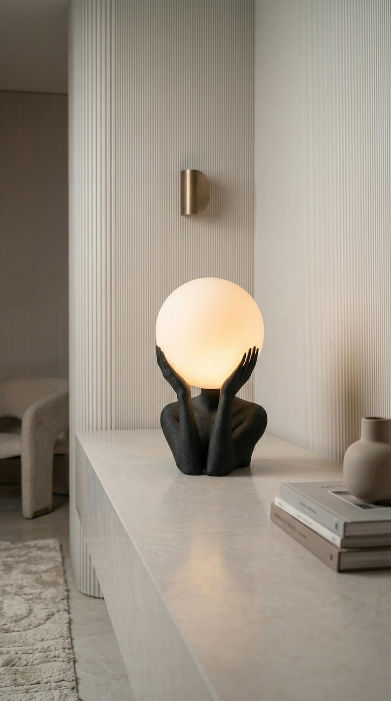 May include: A table lamp featuring a matte black textured sculpture of a torso and hands cradling a large, glowing, spherical white glass shade. The light emits a warm, soft illumination, highlighting the detailed form of the hands and arms.