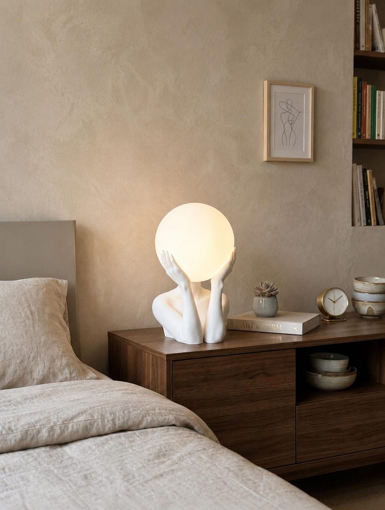 Puede incluir: Una l&aacute;mpara de mesa blanca con una figura humana esculpida desde el pecho, con las manos levantadas para sostener una gran pantalla esf&eacute;rica de cristal blanco que emite una luz suave y c&aacute;lida.