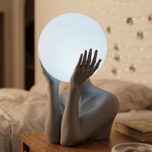 May include: A stone gray sculptural lamp featuring a figure with raised arms holding a glowing white spherical light. The base of the lamp is shaped like a torso and arms, and the text Serenity Sculptural Lamp in Stone gray is visible.
