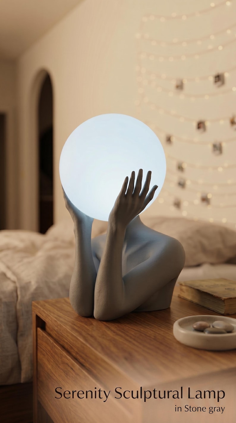 May include: A stone gray sculptural lamp featuring a figure with raised arms holding a glowing white spherical light. The base of the lamp is shaped like a torso and arms, and the text Serenity Sculptural Lamp in Stone gray is visible.