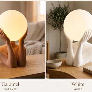 Puede incluir: Una l&aacute;mpara de mesa con un torso humano esculpido y brazos que sostienen una bombilla esf&eacute;rica brillante. La l&aacute;mpara est&aacute; disponible en dos colores: un tono caramelo c&aacute;lido y un acabado blanco mate, ambos emitiendo una luz suave y difusa.