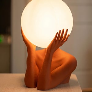 May include: A sculptural lamp in a terracotta finish featuring a torso with arms raised to hold a large, glowing, spherical white light bulb. The base of the lamp is shaped like a human upper body, and the text Serenity Sculptural Lamp in Terracotta is printed at the bottom.
