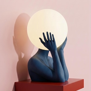 May include: A navy blue sculptural lamp featuring a figure with raised hands holding a glowing white spherical light bulb. The lamp rests on a rectangular red base. The text Serenity Sculptural Lamp in Navy is printed at the bottom of the image.