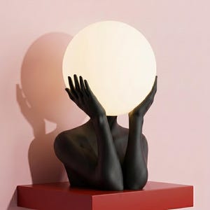 May include: A sculptural lamp featuring a matte black figure with arms raised, holding a large, glowing, spherical white glass shade. The base is a solid red rectangular shelf. The text Serenity Sculptural Lamp in Black is printed below.