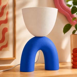 May include: A table lamp featuring a matte white bowl-shaped shade resting atop a vibrant blue arch-shaped base. The lamp has a smooth, solid-colored finish and includes a white power cord extending from the back of the blue arched structure.