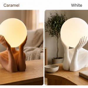 Puede incluir: Dos l&aacute;mparas de mesa que presentan un torso humano y brazos sosteniendo una bombilla esf&eacute;rica brillante. Una l&aacute;mpara es de color caramelo y la otra es blanca. Las l&aacute;mparas est&aacute;n etiquetadas como Caramel y White, mostrando las opciones.