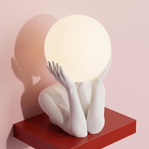 May include: A white sculptural lamp featuring a pair of hands cradling a glowing spherical light bulb. The sculpture is mounted on a rectangular red shelf. The text "Serenity Sculptural Lamp in White" is printed at the bottom of the image.