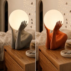Puede incluir: Una l&aacute;mpara de mesa con una figura esculpida cuyos brazos levantados sostienen una bombilla esf&eacute;rica blanca brillante. La base est&aacute; disponible en dos acabados mate: un gris piedra neutro y un color terracota c&aacute;lido.