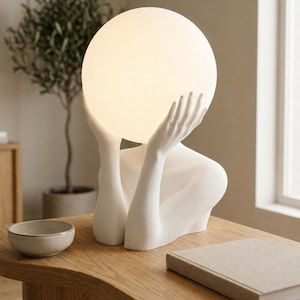 Puede incluir: Una l&aacute;mpara de mesa blanca con una figura esculpida cuyos brazos levantados sostienen una gran bombilla esf&eacute;rica brillante. La base es un busto blanco mate que muestra el torso, los hombros y las manos que sostienen la esfera iluminada.