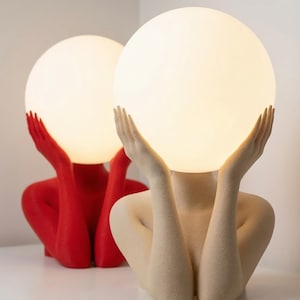 May include: Two table lamps featuring a human-like torso and arms holding a large, glowing, spherical white light bulb. One lamp is finished in a solid matte red, while the other is finished in a neutral, textured beige color.