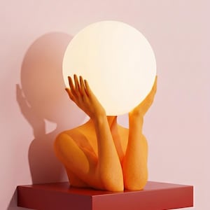 May include: A sculptural lamp featuring an orange textured figure with arms raised, holding a large, glowing, spherical white light bulb. The lamp is mounted on a rectangular red base and displays the text Serenity Sculptural Lamp in Orange.