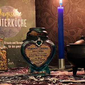Könnte beinhalten: Eine herzförmige Flasche in Türkis mit der Aufschrift "Verdant Desire", einem Korken und einem grünen Herzanhänger. Eine blaue Kerze brennt neben einem kleinen schwarzen Kessel. Ein Buch mit deutschem Text im Hintergrund.