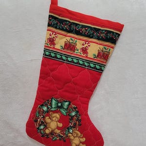 Peut inclure: Une chaussette de Noël rouge matelassée avec une bande supérieure ornée de motifs de sucres d'orge et de tambours. Le corps présente une couronne imprimée avec un nœud vert et deux oursons marron clair. Le tissu a un motif de grille cousu blanc.