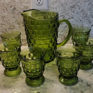 May include: A green glass pitcher with a handle and six matching glasses. The pitcher and glasses have a textured, geometric pattern. The glassware is arranged on a light-colored surface.