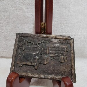 May include: A rectangular metal belt buckle featuring a relief design of a semi-truck hauling a trailer loaded with logs. The buckle has a textured, wood-grain border and a dark, antiqued bronze finish, depicting a detailed logging vehicle scene.