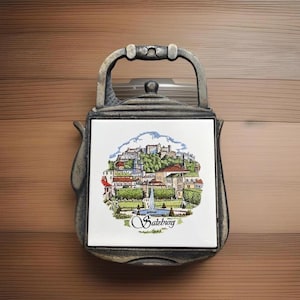 May include: A decorative metal trivet in the shape of a teapot, featuring a ceramic tile with a colorful illustration of Salzburg, Austria. The tile depicts buildings, trees, and a fountain, with the word "Salzburg" in script.