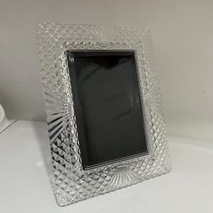 May include: A clear, rectangular picture frame with a diamond-patterned border and a silver-colored inner frame. The frame has a decorative fan-like design at the top and bottom. The frame is designed to hold a rectangular photo.
