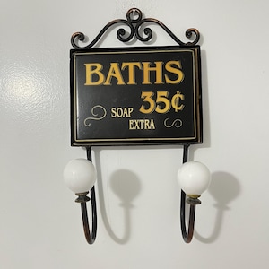 May include: A black metal wall-mounted sign with gold lettering that reads BATHS 35¢ SOAP EXTRA. The sign features a decorative scrollwork top and two downward-curving hooks, each finished with a smooth, round white ceramic knob at the end.