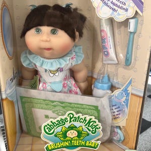 May include: A Cabbage Patch Kids "Brushin' Teeth Baby" doll in its original packaging. The doll has brown hair, green eyes, and is wearing a floral dress. Accessories include a toothbrush, toothpaste, and a baby bottle. The packaging includes the text "Brushin' Teeth Baby".