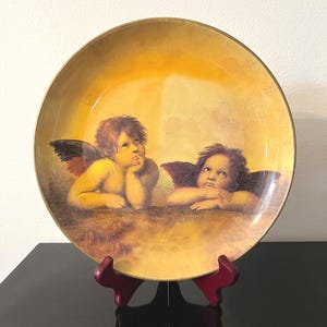 May include: A round decorative plate featuring a warm yellow and gold gradient background. The center displays a detailed illustration of two cherubs with dark wings, resting their chins on their hands while looking upward in a contemplative pose.