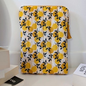 Peut inclure: Une housse d'ordinateur portable matelassée avec une fermeture éclair. Le tissu présente un motif répétitif de citrons jaunes et de fleurs blanches sur un fond crème.