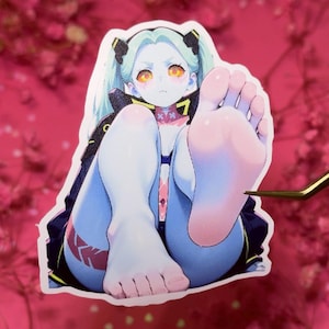 May include: A sticker featuring an anime-style character with pale blue skin, yellow eyes, and light blue hair in pigtails. The character is seated, with legs extended and feet facing forward. The sticker has a pink and red color scheme.