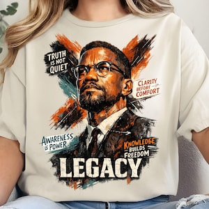 May include: A cream-coloured t-shirt with a portrait of a man wearing glasses, and the word "LEGACY" in bold letters. The design includes text that reads "TRUTH IS NOT QUIET", "AWARENESS IS POWER", "CLARITY BEFORE COMFORT", and "KNOWLEDGE BUILDS FREEDOM".