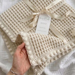 Può includere: Coperta per neonati color crema a maglia a nido d'ape, legata con un nastro color crema. La coperta presenta un motivo quadrato strutturato e un bordo decorativo con piccole palline rotonde. Un'etichetta è attaccata al nastro.