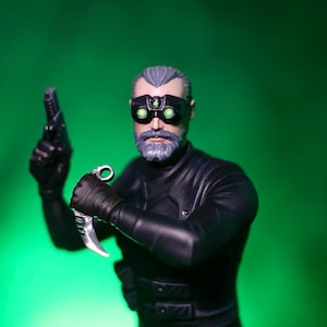 Op de afbeelding: Een beeldje van een man in een zwart tactisch pak, met een pistool en een gebogen mes. Hij draagt zwarte handschoenen en een nachtzichtmasker met groene lenzen. De figuur heeft een grijze baard en haar, tegen een groene achtergrond.