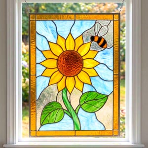 May include: Stained glass window panel depicting a sunflower with yellow petals and a brown centre, a green stem and leaves, and a bee. The background is light blue with a gold border. The panel is hanging in a white window frame.