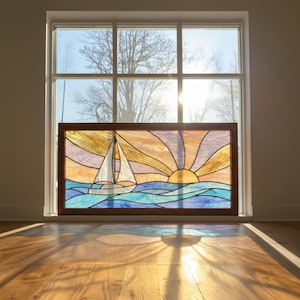 以下が含まれることがあります： 夕焼けを背景に、海に浮かぶヨットを描いたステンドグラスパネル。木製のフレームが特徴で、窓の前に飾られています。太陽がガラスを通して輝いています。